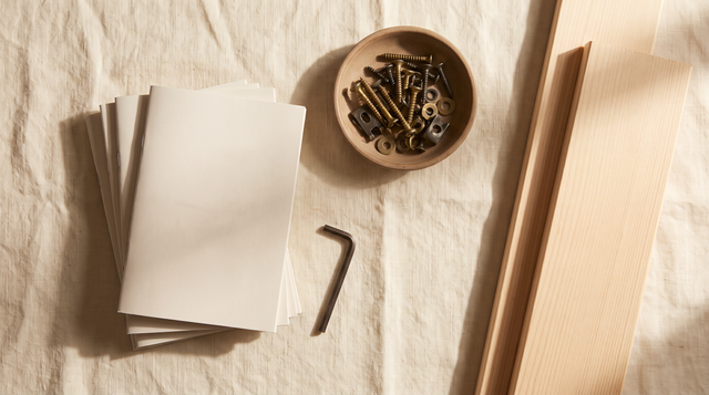 Overhead view of a stack of sauna assembly manuals beside a hex key, a dish of assembly hardware and pale hemlock wall panels