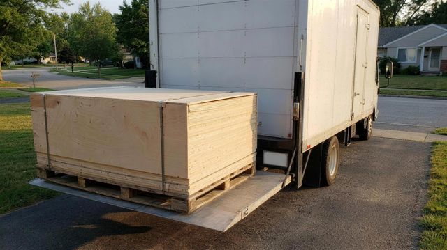 Flat-packed sauna crate on a pallet at a freight truck liftgate in a driveway